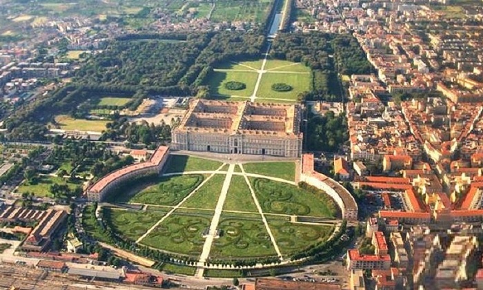 Città-e-Reggia-di-Caserta - NCC Napoli Aerial view of a grand European palace with circular formal gardens and radiating paths surrounding the central lawn.